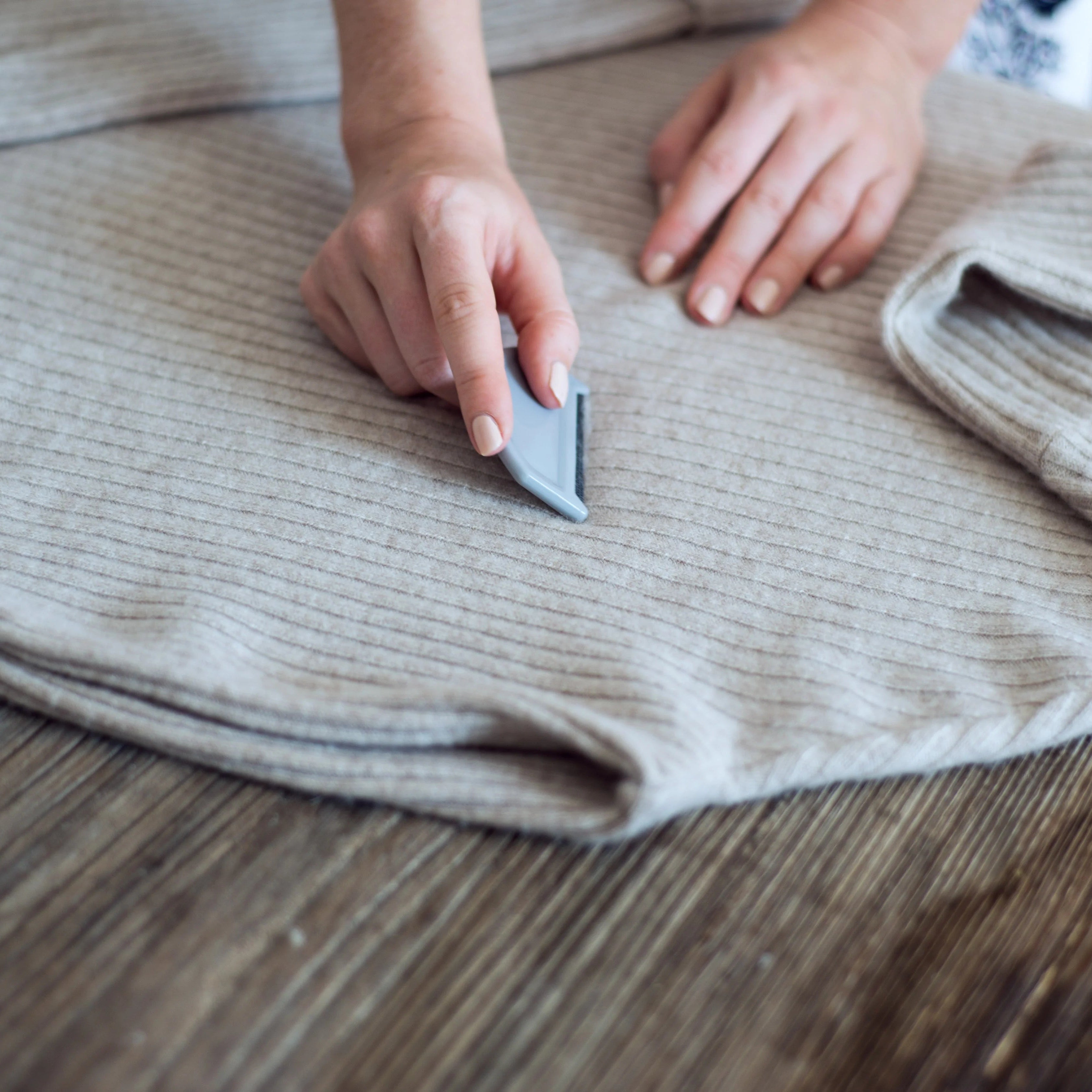 Person using a small tool on a folded beige sweater on a wooden surface